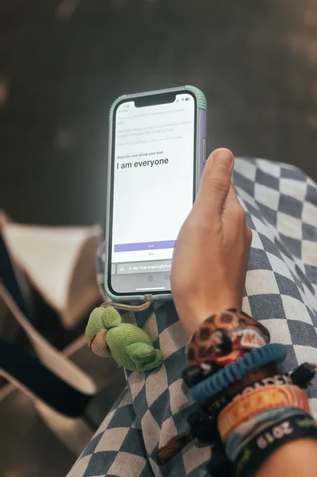 An image of right hand holding phone on lap. On screen it has a small text that blur and the readable text said “ Read this text and stomp your feet. I am everyone”