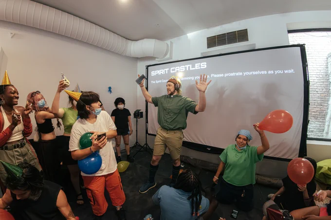 A person wearing a green shirt with a light on the head put the arms up in the air in front of the projection screen. Surrounding by many people holding balloons in a party mood