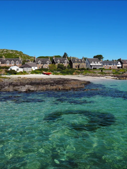 Image descriptiomn: crystal clear blue water leading to a small beach lined with stone houses. 
