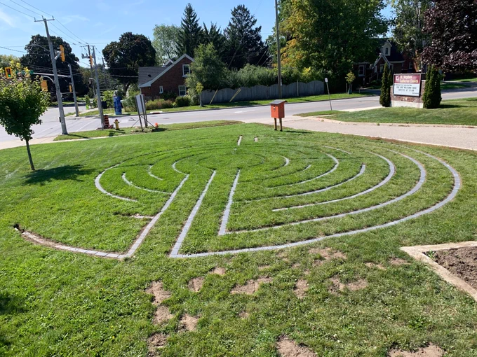 A circular labyrinth, made of paving stones set into a lawn, is being built. The area to the right is finished, the part on the left needs more work. There is an orange little library at the far side of the labyrinth.