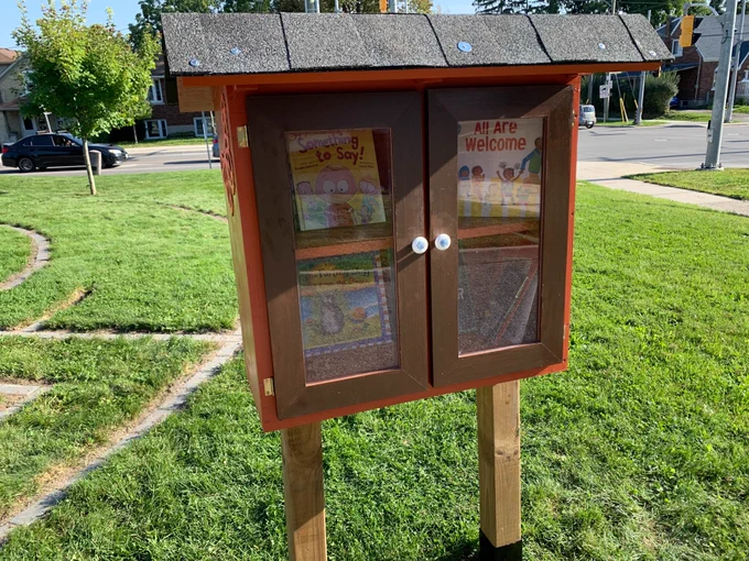 An orange little library with a few children's books in it. One title we can see is "All Are Welcome."