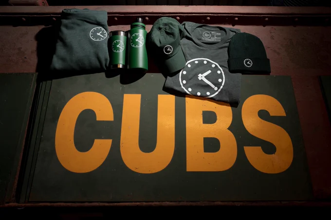 An image promoting the Obvious Shirts x Cubs giveaway. The photo shows a display of Obvious Shirts 1:20 clock hoodies, t-shirts, hats, and water bottles.