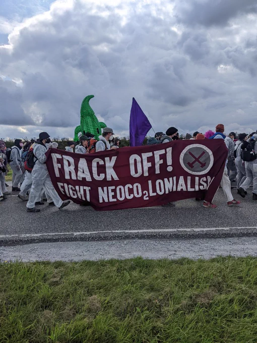 Protest rally on route, activists in overalls carrying inflatable dinosaurs, flags and a banner saying 'Frack off! Fight NeoCO2lonialism'