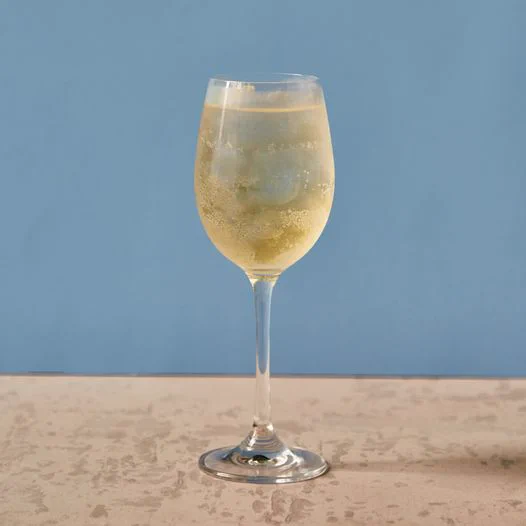 A light yellow, nearly see-through cocktail with ice in a glass, with a blue background.
