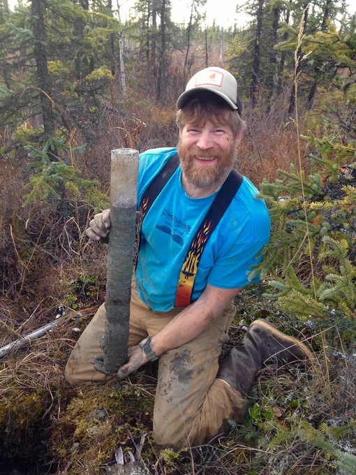 Photograph of a man wearing a baseball hat and a blue t shirt with overalls holding up a permafrost core while kneeling on the ground in a forest.