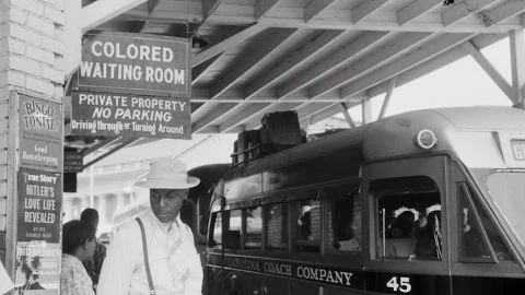 A photo of train station in the 1940s. A young Black man wearing a white suit and hat is standing under a sign which reads "Colored Waiting Room." 