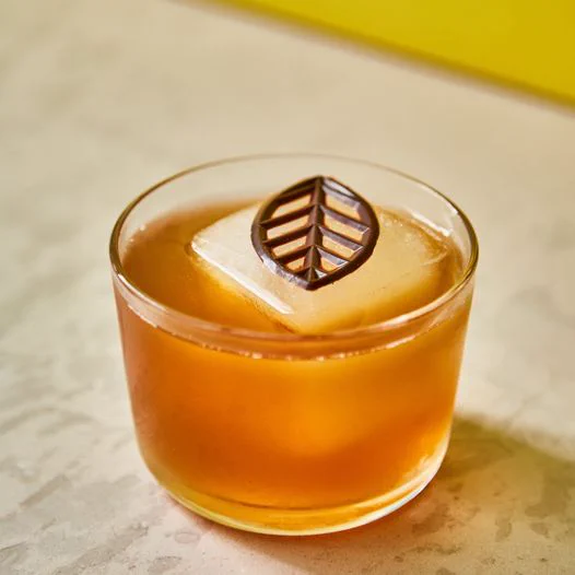 A deep yellow cocktail in a short glass, with a big ice cube and a chocolate leaf garnish