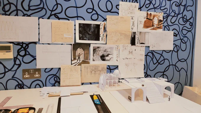 Studio space for Designer and Maker resident (Rimu) Tung K H, including paper maquettes and tools on the desk See https://bit.ly/TD_Designer and artwork pinned to the wall, decorated with a blue squiggly design by Nicole-Rose Munhawa.