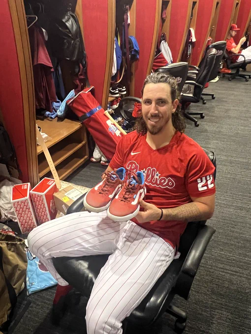 Photo of Michael Lorenzen sitting in the Phillies clubhouse.  He is holding a pair of dark red Vans cleats.  He is wearing a red Phillies t-shirt with red and white pinstripe baseball pants.