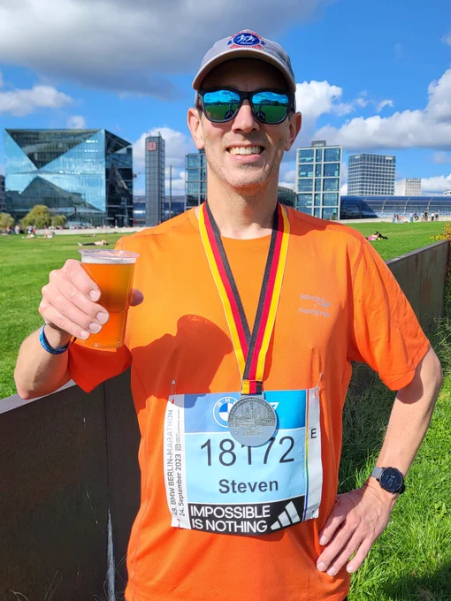 A man smiles while wearing an orange running shirt and tan running hat with a Berlin Marathon medal around his neck. In the background is the Berlin Central Railroad Station.