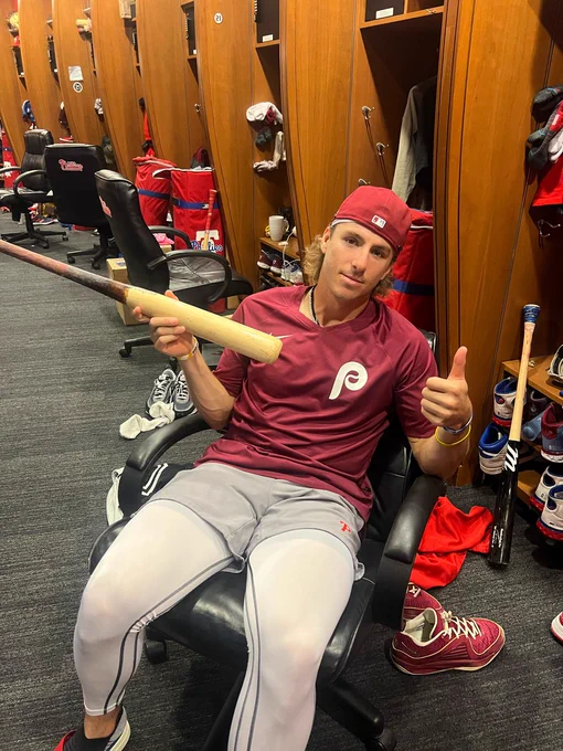 Photo of Bryson Stott sitting in the Phillies clubhouse holding one of his bats.  He is giving a thumbs up.  Stott is wearing a dark red Phillies t-shirt, gray Phillies shorts with white pants underneath and a backwards red hat.