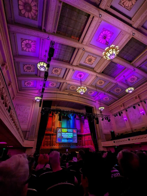 Inside a packed Melbourne Town Hall, lights illuminate the historic grand organ in the colours of the rainbow pride flag — red, orange, yellow, green, blue, and purple. Elsewhere, pink and purple lights reflect off the ornate town hall ceiling. The screen says: "Welcome to the Melbourne Queer Film Festival twenty twenty three Program Launch."