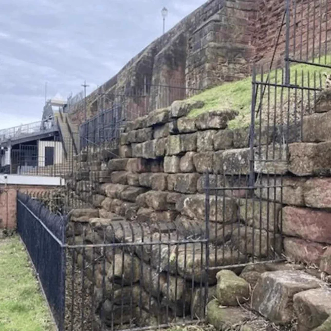A photo of some ancient stone work. It appears to be a stepped wall in sandstone, that has done black metal railings around it. It is at the base of the city wall.  

Image: © Chris Clayton.