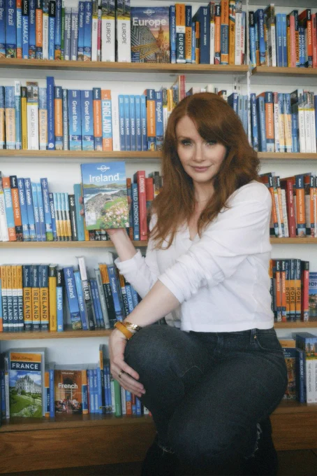 Wearing a white blouse and blue jeans, BDH stands in front of a bookcase with shelves and shelves of miscellaneous travel guides. She holds the Lonely Planet guide for Ireland by her face, absolutely ecstatic about returning to Ireland after 30 years!!