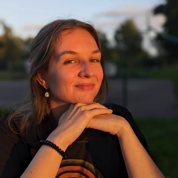 Photograph of a woman sitting facing the sun and the camera leaning on both hands with a blurred background consisting of trees, ground and sky.