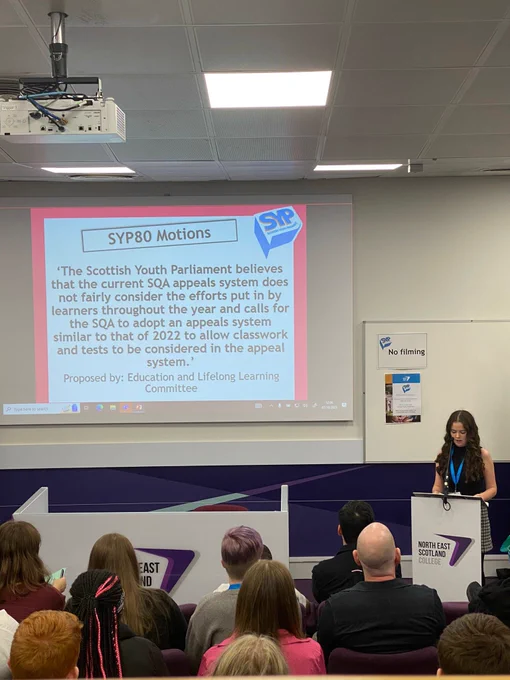 A big screen reading “SYP80 Motions ‘The Scottish Youth Parliament believes that the current SQA appeals system does not fairly consider the efforts put in by learners throughout the year and calls for the SQA to adopt an appeals system similar to that of 2022 to allow class work and tests to be considered in the appeal system.’ Proposed by, Education and Lifelong Learning Committee” with me standing at the front proposing the motion.