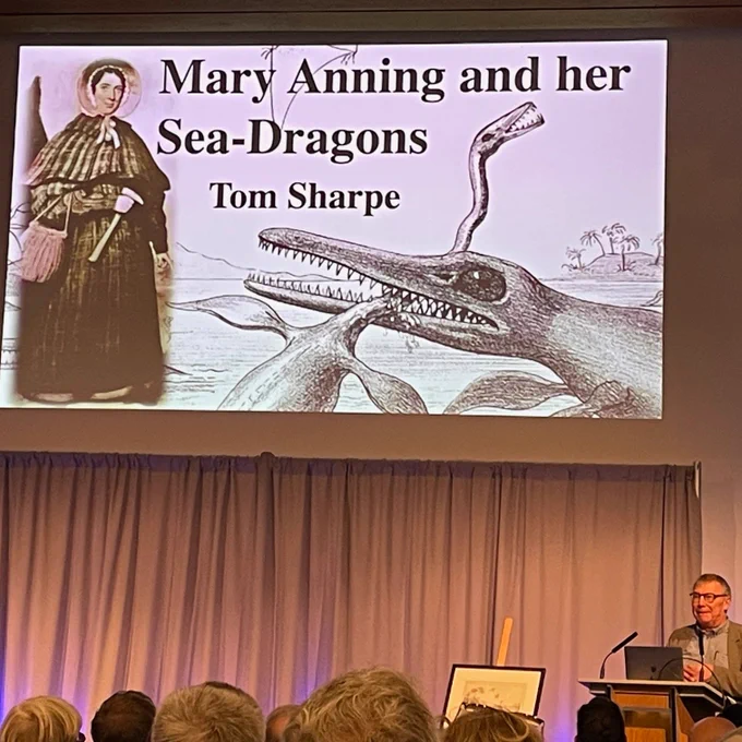 A man is facing an audience of people standing behind a lectern giving a presentation. Above the man is a large screen showing a presentation slide with a picture of Mary Anning and a drawing of two extinct marine reptiles. One of the extinct reptiles has a long jaw with many long teeth similar in appearance to a modern day crocodile. The other extinct reptile has a very long thin neck and two front flippers and its neck is being bitten by the other reptile.