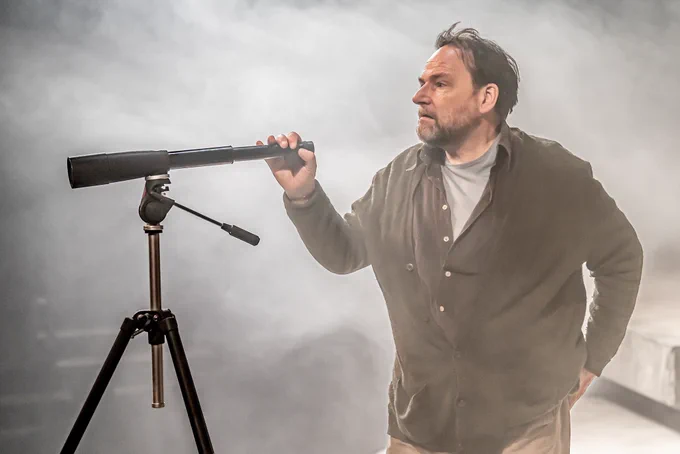 Peter Forbes (Robin) in On The Beach. Photo by Marc Brenner. Peter wears a cardigan and stands in white haze, one hand holding the end of a telescope mounted on a tripod.