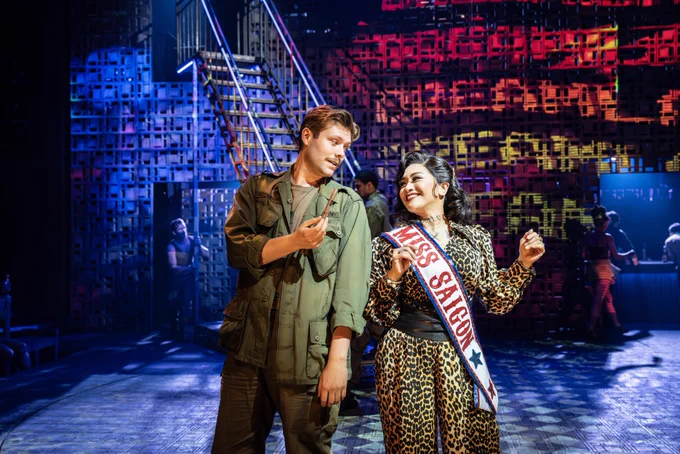 Shane O’Riordan (John) and Joanna Ampil (The Engineer) in Miss Saigon. Photo by Johan Persson. Shane wears a green army uniform and leans over to offer Joanna a cigar. Joanna smiles and looks at Shane. Joanna wears a sash which reads ‘Miss Saigon’.
