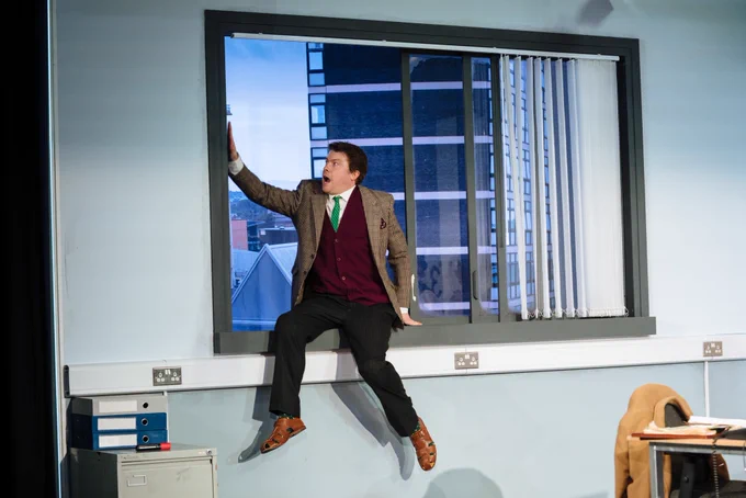 Daniel Rigby (The Maniac) in Accidental Death of an Anarchist. Photo by Helen Murray. Daniel sits on a window ledge, their feet dangling. They hold the frame with each hand and look out at the drop below.