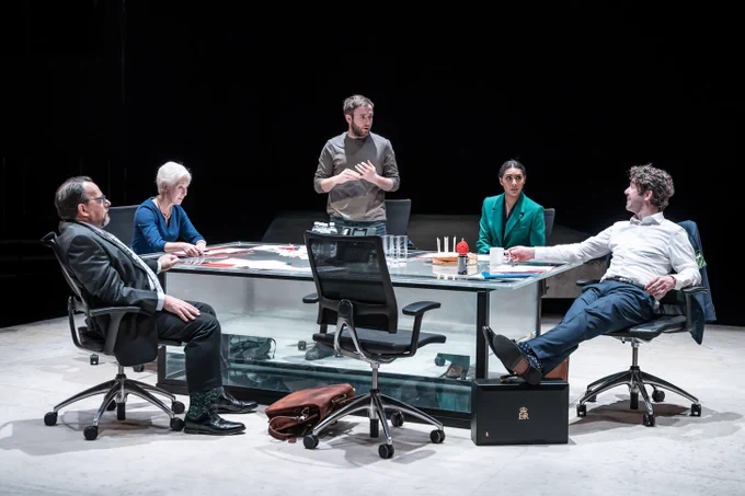 The company of Resilience. Photo by Marc Brenner. Four people of varying age, gender and ethnicity wear formal office-wear and sit around a desk. Joe Bannister stands in the centre of them and wears a casual t-shirt. They look worried and wring their hands as they speak to the seated group.