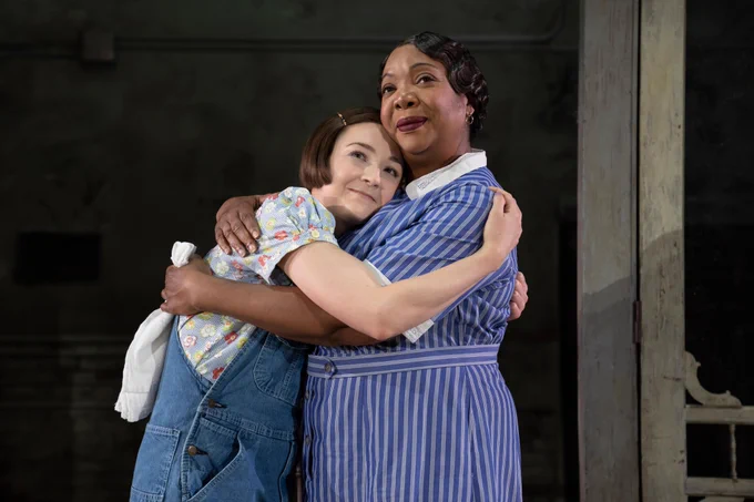 Two actors on a stage, hugging each other. They are both smiling. 