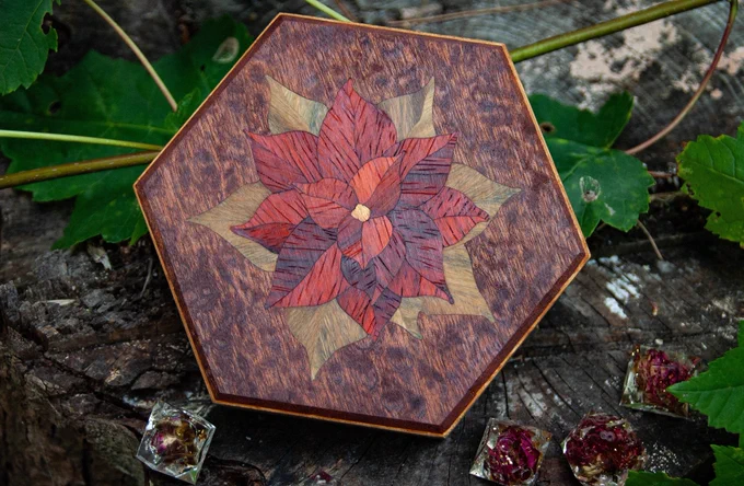 Hexagonal marquetry dice box with the likeness of a Poinsettia plant depicted. It is resting on a stump with maple leaves and dice acting as set dressing.
