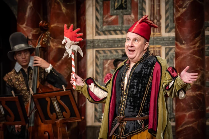 The cast of Twelfth Night: For One Night Only onstage in the Globe Theatre (2023) - one dressed as a fool gesturing, with a musician behind.