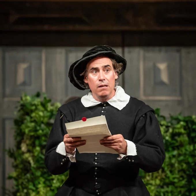 The cast of Twelfth Night: For One Night Only onstage in the Globe Theatre (2023) - one cast member reading a letter and looking out into the audience pensively, with a green bush behind them.