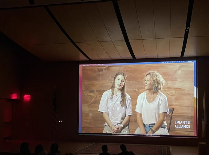 People sit in a theater looking at a screen where two people are talking
