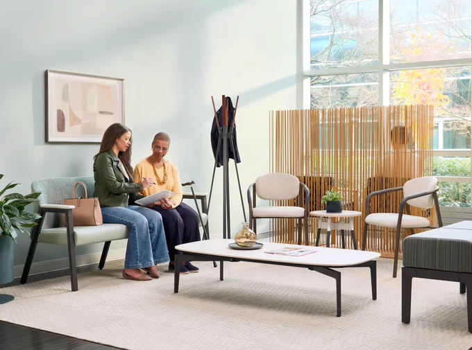Two women sit in a modern healthcare waiting room featuring Steelcase Health Radia chairs and benches and the Steelcase Health Embold table.