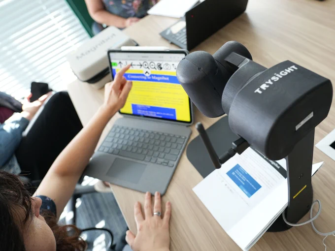 Finger points to a laptop screen with magnified material on it. The MagniBot sits beside. Highlighting colours on screen are high contrast, yellow and Blue.
