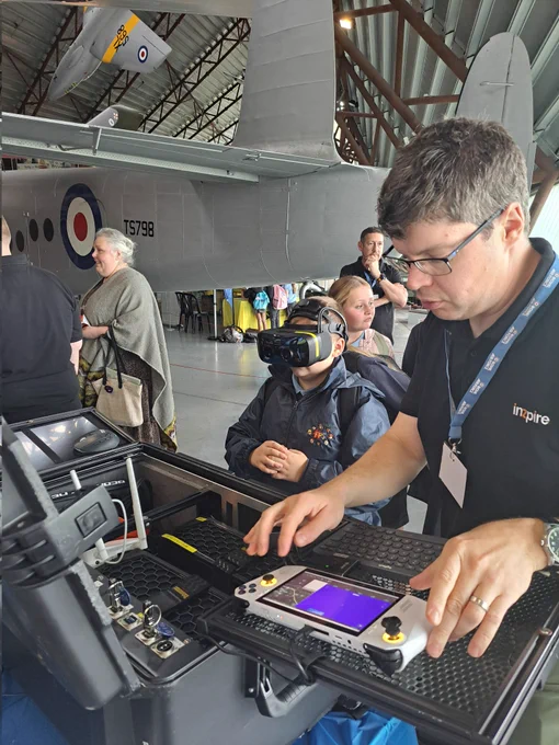 Man with glasses demonstrating using a games controller to a child with a VR headset on.