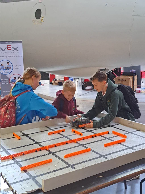 Three children guiding a robot on a board, with a sign that reads 'VEX Robotics' in the background.