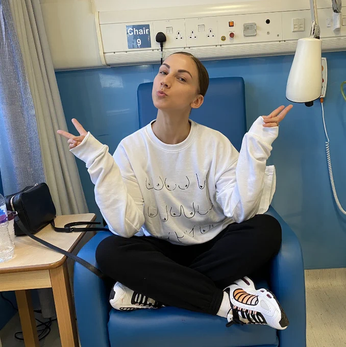 Becksy is sitting in a chair in hospital wearing a jumper with boobs on it. She is posing for the camera.