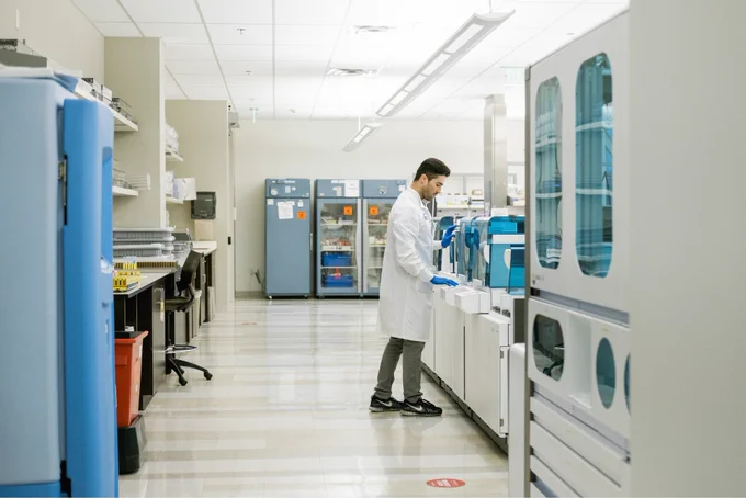 Person with short dark hair wearing a white lab coat stands at a science bench in a lab.