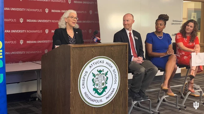 Interim Chancellor Murdoch-Kinch (left) stands behind a podium and speaks at Crispus Attucks High School, a group of people (right) sitting and watching from the side.