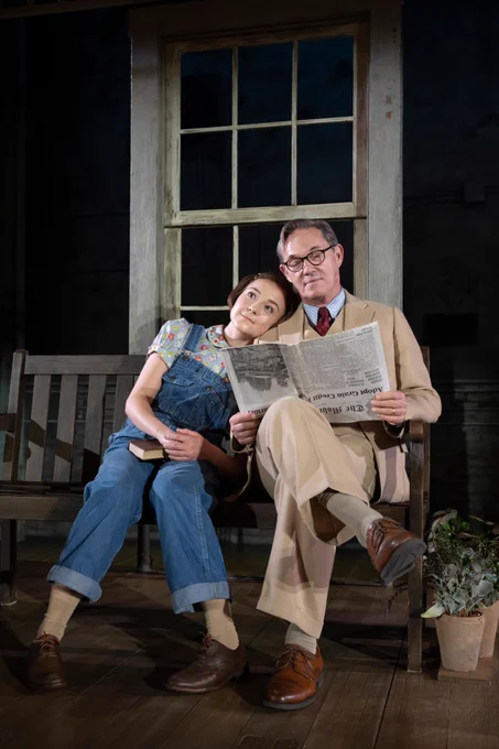 2 actors sitting on a bench together. The person on the left of the photo is wearing circular glasses and pointing to the person on the right while speaking to them. 