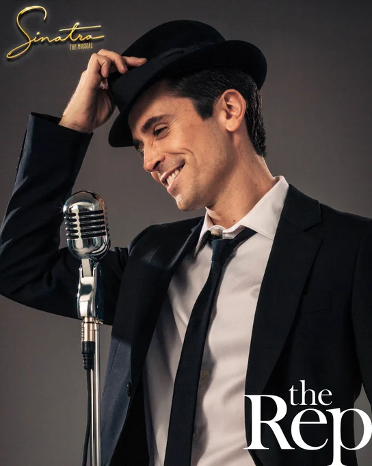 A young man wearing a trilby hat stands smiling in front of a retro microphone. 