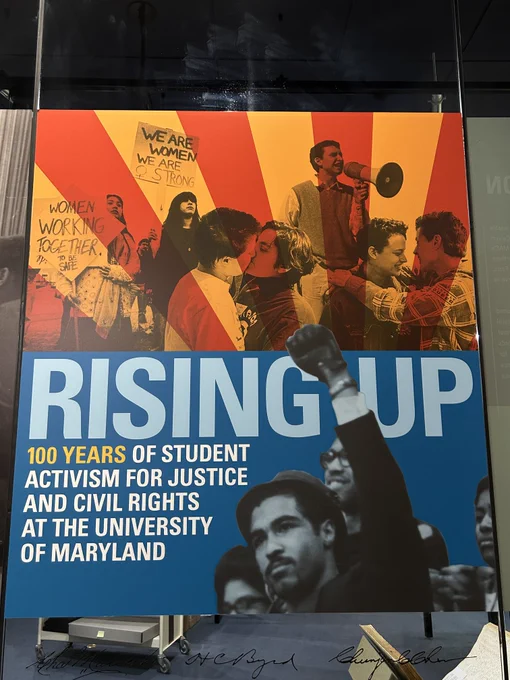 Color photograph of the exhibition sign. The top half of the sign is a photo of two women at a march, holding signs that read "we are women / we are strong / women working together to be safe". Beside them are photos of gay couples kissing and holding each other at a rally. There is a yellow and orange filter on these photos. The bottom half of the sign is a photo of a man at a rally holding up his fist in the air. The text across the sign reads "Rising Up / 100 years of Student Activism for Justice and Civil Rights at the University of Maryland"