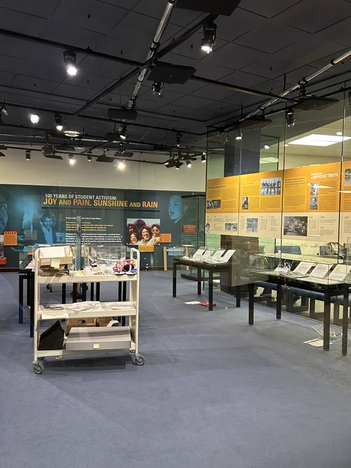 Color photo of the inside of the gallery. There are glass cases along the wall and some wall panels with photos and text hung up. There is a white cart in the foreground of the photo holding supplies like boxes, scissors, paper, etc.