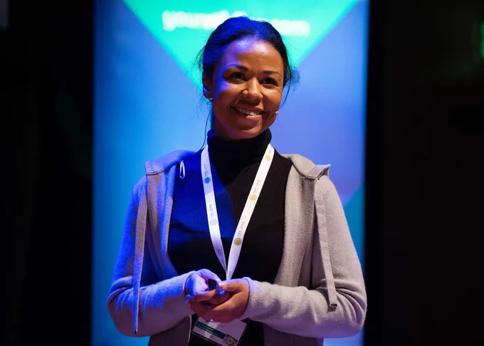 A woman stands on stage in a black turtle neck and grey hoodie. She smiles at the audience.