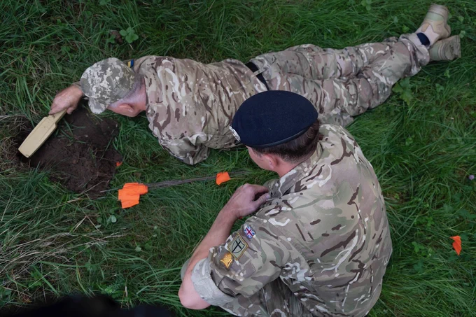 A soldier from the Ukrainian Armed Forces lies flat on grassed ground with a handheld electronic device for mine detection in his right hand. He is using the device over a shallow hole in the ground. Beside him, a Royal Engineer is on his haunches observing the training. Both are wearing green/brown/grey camouflaged uniforms. The Ukrainian soldier has a cap and the British soldier a dark beret.