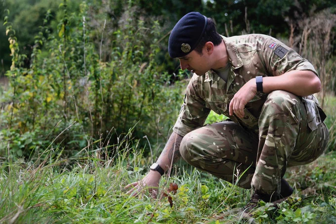 On the right of the image a Royal Engineer soldier is on his haunches in an area of grassed/shrubbed area. He is wearing a green/black/grey/khaki camouglaged uniform and a dark beret. His right hand is on the ground gently looking for signs of unexploded devices.