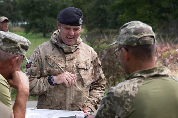 Three soldiers can be seen - two Ukrainian soldiers in the foreground with their backs to the camera and one, a British Army. Royal Engineer in the centre looking towards it and is smiling. The British soldier is wearing a green/brown/black/khaki camouflaged uniform jacket and a dark beret. In his hand he holds a thin stick to point towards a map of the area where the training is being carried out. The Ukrainian soldiers are wearing camouflaged caps and green shirts.