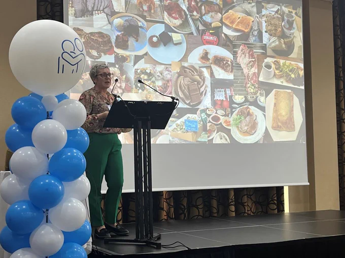 A woman is standing behind a podium with a large screen behind her which is full of photographs of food. 
