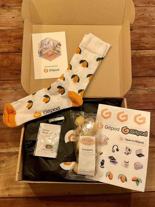 A photo of an opened surprise box from Gitpod featuring socks, kumquats sweets, stickers, a bottle opener and a webcam cover.