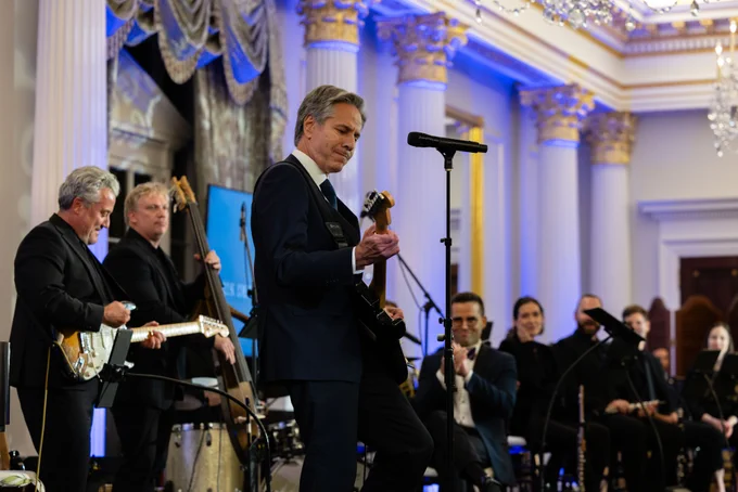 Secretary of State Blinken plays the guitar during a launch event for the Global Diplomacy Music Initiative at the Department of State. A crowd and band watches in the background from an ornate and blue-tinted diplomatic event space.