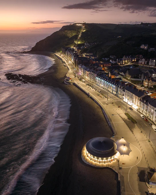 An aerial photograph of north beach in Aberystwyth in warm dusk light showing the bandstand in the bottom of the shot, the promenade and Constitution Hill. The sea is blurred by long exposure.