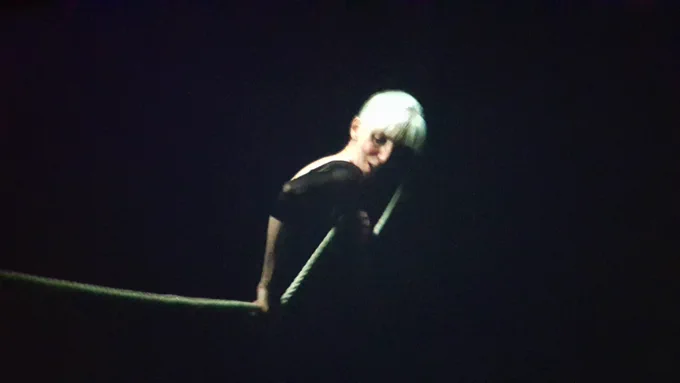 Photo of an older dancer in a dark space. They are wearing black and holding a rope.